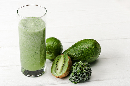 green smoothie in glass near kiwi, broccoli, avocado and lime on white wooden surface