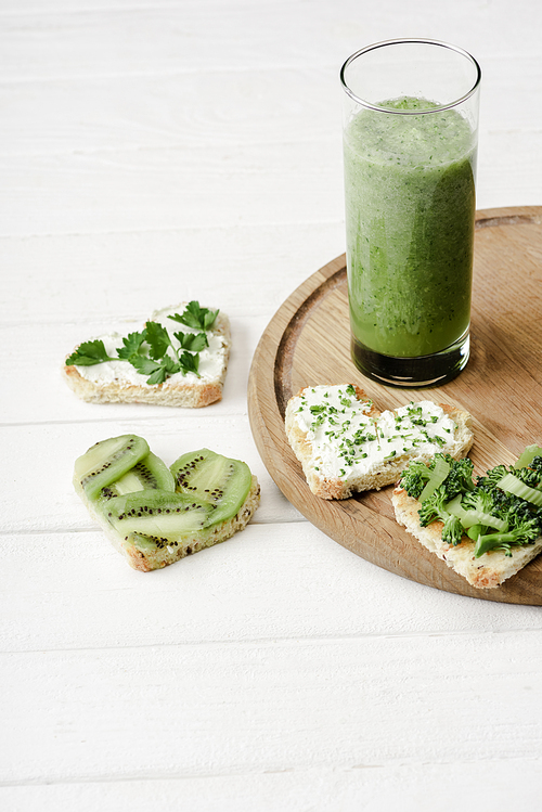 green smoothie and heart shaped canape with creamy cheese, broccoli, microgreen, parsley and kiwi on white wooden surface