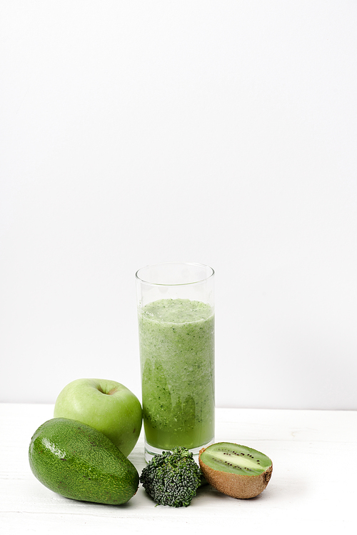 green smoothie in glass near kiwi, broccoli, avocado and apple on white wooden surface