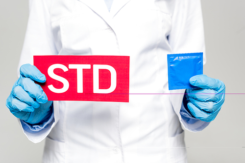 cropped view of doctor in white coat and latex gloves holding paper with std lettering and condom isolated on grey