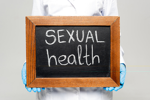 cropped view of doctor in white coat and latex gloves holding chalkboard with sexual health lettering isolated on grey