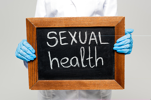 cropped view of doctor in latex gloves holding chalkboard with sexual health lettering isolated on grey