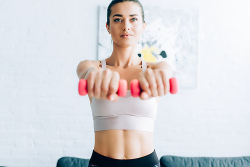 Selective focus of brunette sportswoman working out with pink dumbbells at home