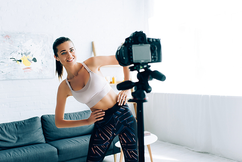 Selective focus of sportswoman training near digital camera on tripod in living room