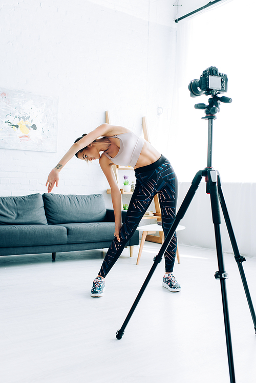 Selective focus of young sportswoman stretching near digital camera in living room