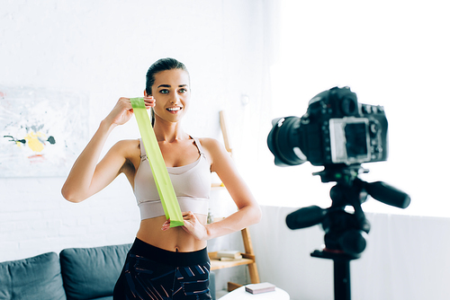 Selective focus of sportswoman holding resistance band near digital camera on tripod at home