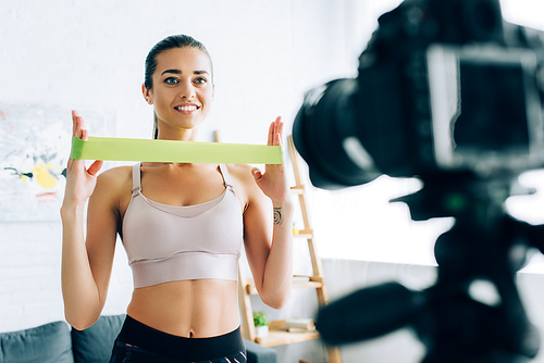Selective focus of fit sportswoman holding resistance band near digital camera in living room
