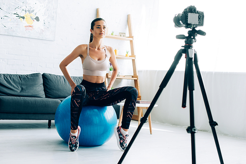 Selective focus of sportswoman sitting on fitness ball near digital camera on tripod at home