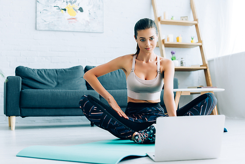 Selective focus of brunette woman in sportswear watching online training on laptop on fitness mat