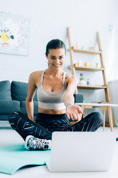 Selective focus of young sportswoman pointing with hand at laptop while sitting on fitness mat