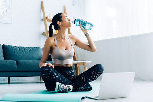 Selective focus of sportswoman drinking water while sitting on fitness mat near laptop