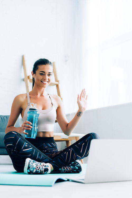 Selective focus of sportswoman waving hand while holding sports bottle near laptop at home