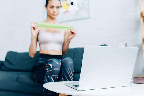 Selective focus of laptop on coffee table and sportswoman holding resistance band on couch