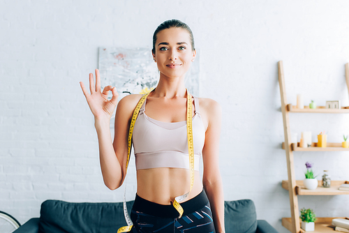 Brunette sportswoman with measuring tape around neck showing okay gesture at home