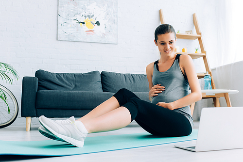 Selective focus of pregnant sportswoman looking at laptop while sitting on fitness mat at home