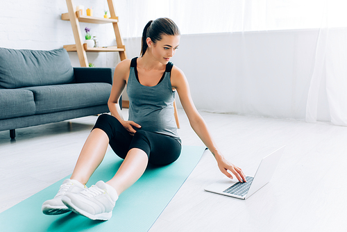 Selective focus of pregnant sportswoman using laptop on fitness mat at home