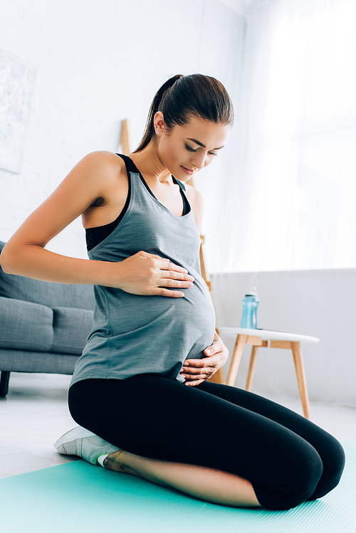 Pregnant sportswoman touching belly while sitting on fitness mat in living room