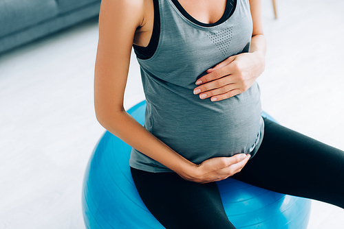 Cropped view of pregnant woman in sportswear touching belly on fitness ball at home