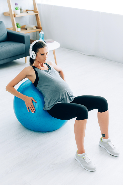 Pregnant sportswoman in headphones training with fitness ball at home