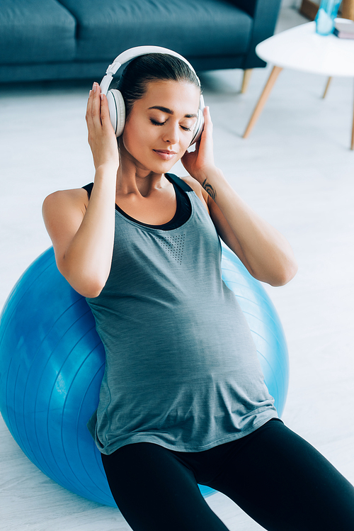 Young pregnant woman listening music in headphones while exercising with fitness ball at home