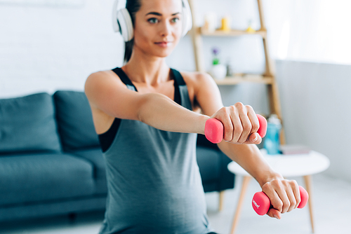 Selective focus of pregnant woman in headphones training with pink dumbbells at home