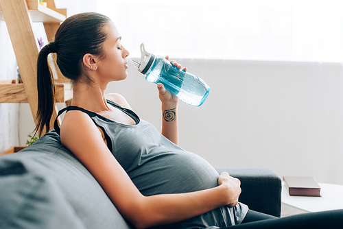 Selective focus of pregnant sportswoman touching belly and holding sports bottle on couch
