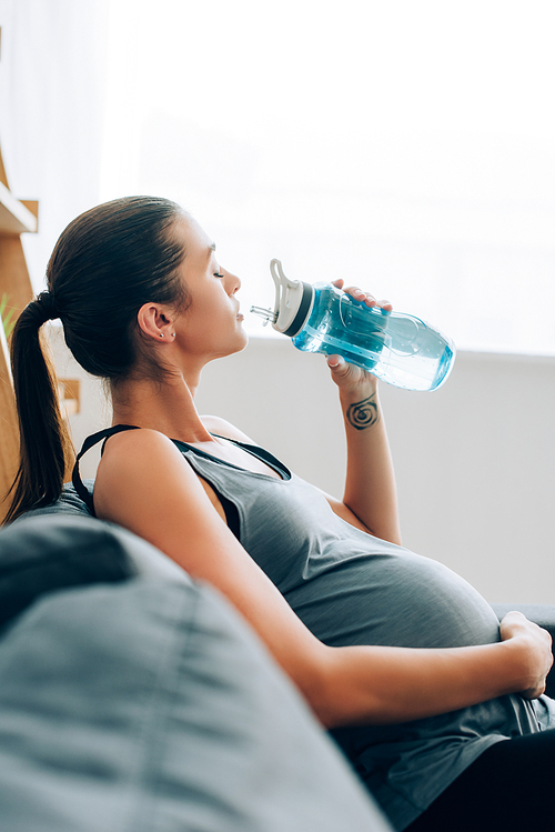 Selective focus of pregnant sportswoman drinking water while sitting on sofa at home