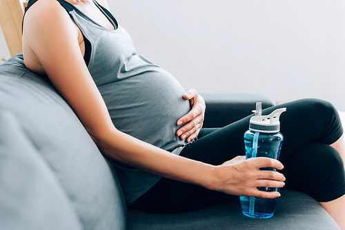 Cropped view of pregnant sportswoman holding sports bottle and touching belly on sofa