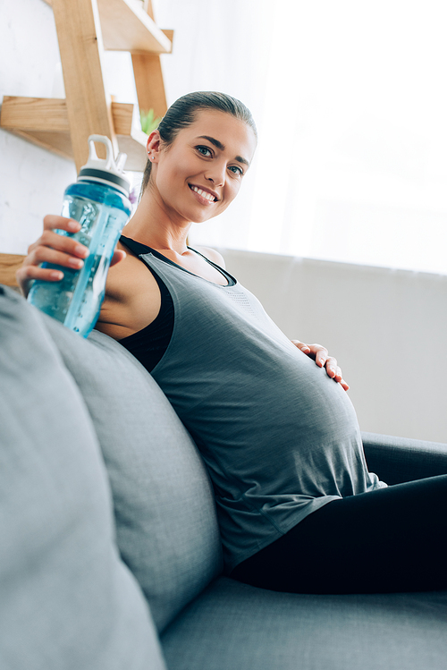 Selective focus of pregnant woman in sportswear holding sports bottle and  in living room