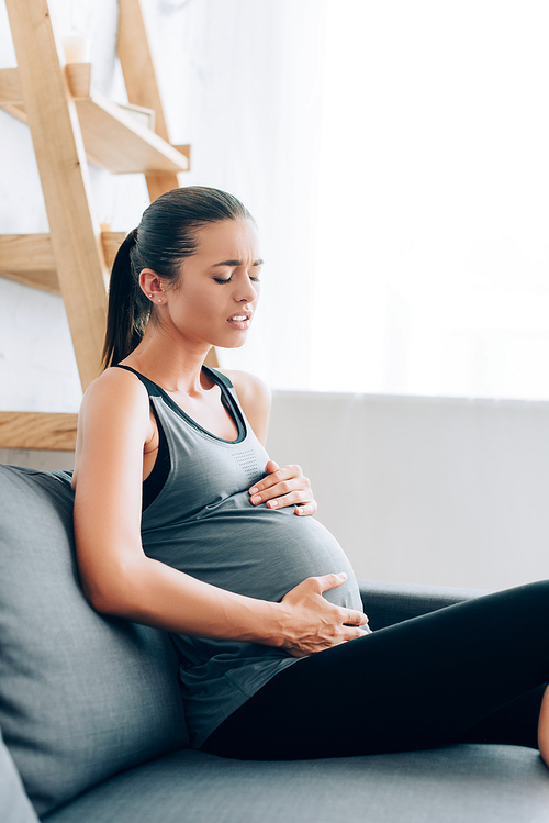 Selective focus of young pregnant sportswoman suffering from hurt on couch at home