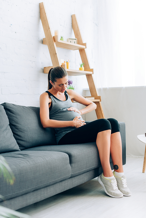Selective focus of pregnant sportswoman feeling pain and touching belly on couch