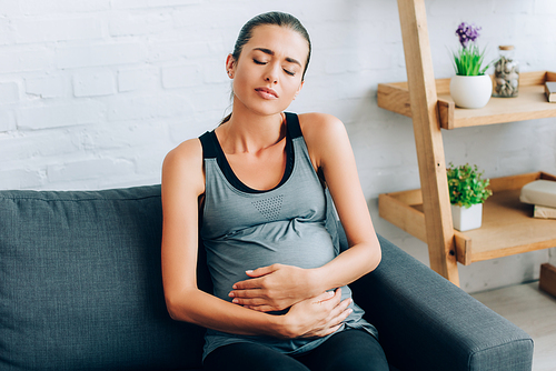 Tired sportswoman embracing belly while sitting on couch at home