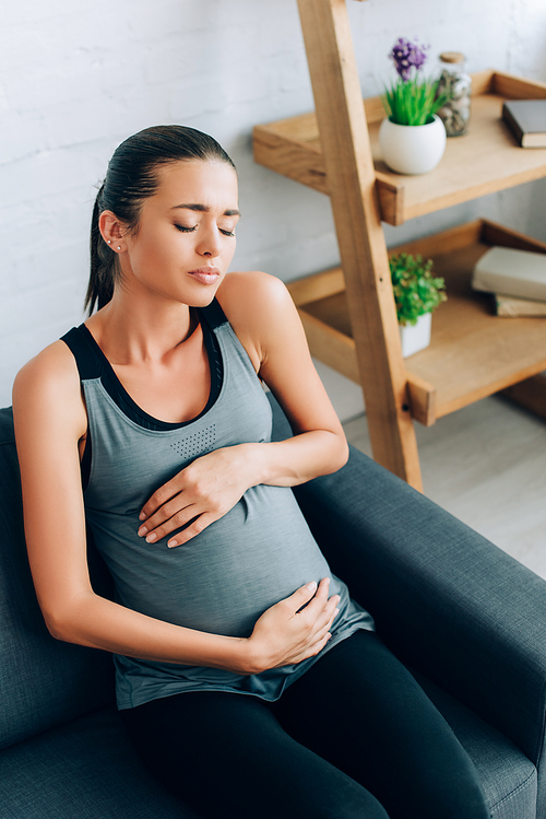 Pregnant sportswoman feeling pain and embracing belly on couch