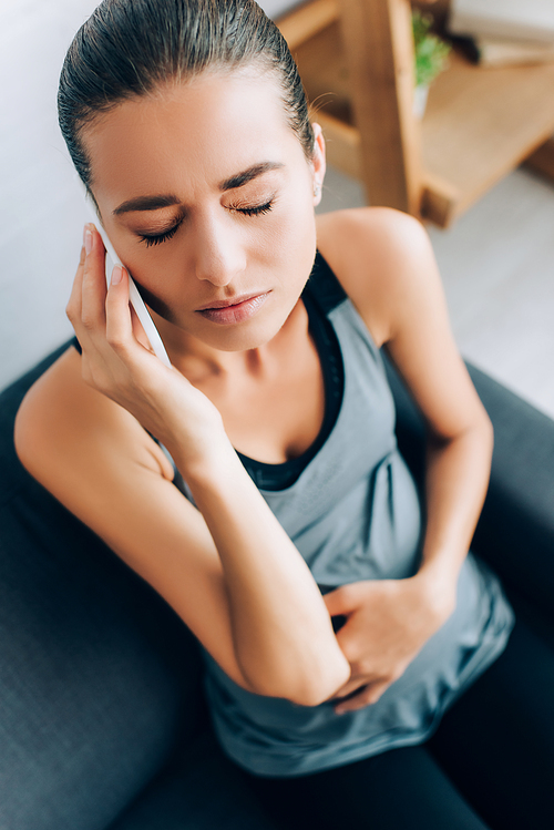 Selective focus of pregnant woman in sportswear talking on smartphone while feeling pain on sofa