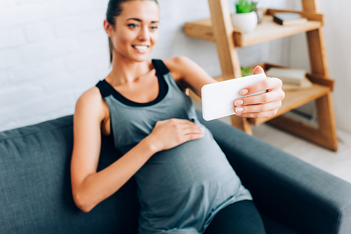 Selective focus of pregnant sportswoman touching belly while taking selfie with smartphone on couch