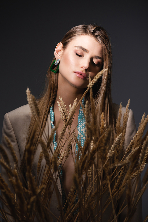 young woman with closed eyes standing near wheat spikelets on dark grey background