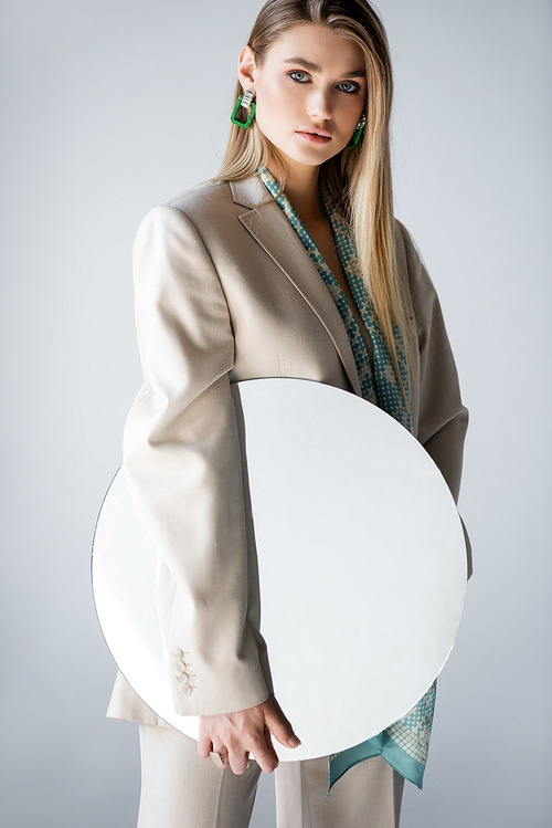 stylish woman in suit holding round mirror while  on grey