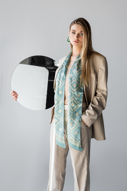 young woman in suit holding round mirror and standing with hand in pocket on grey