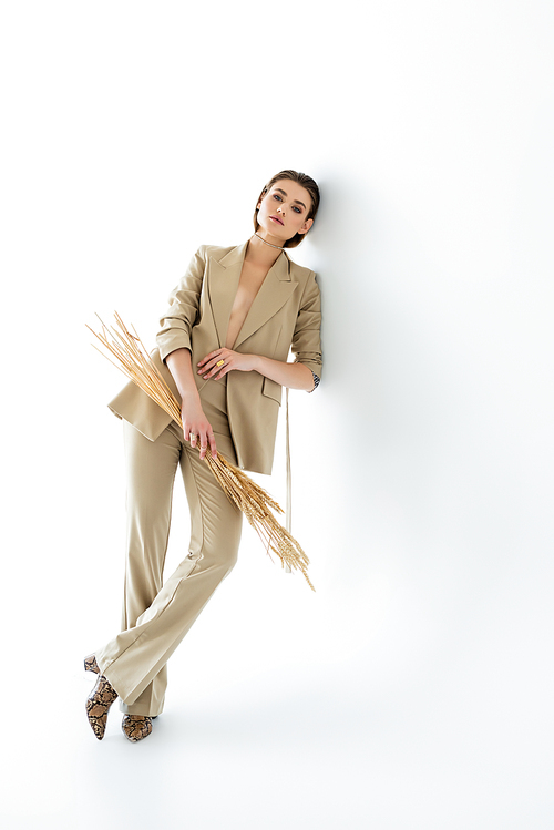 full length of young woman in beige suit posing while holding wheat on white