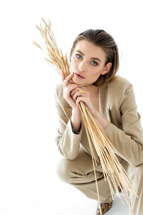 stylish young woman in beige suit sitting while holding wheat on white