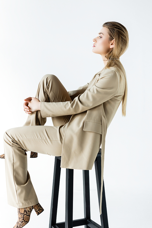 profile of young model in suit poising while sitting on stool on white background