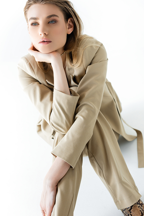 stylish and dreamy model in beige suit sitting on white