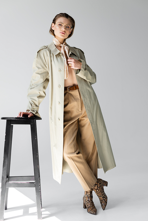 full length of young woman in glasses, trench coat and scarf leaning on stool while posing on grey