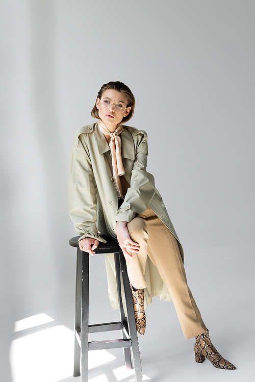 full length of young woman in trench coat and scarf sitting on stool while posing on grey
