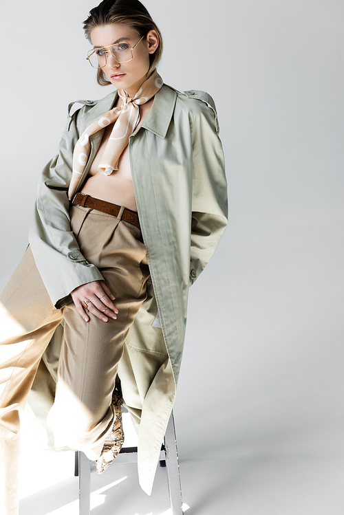trendy young woman in trench coat and scarf sitting on stool while posing on grey