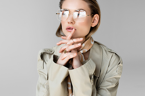 trendy woman in glasses, trench coat and scarf posing on grey
