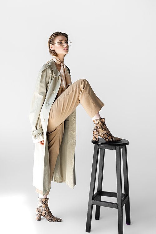 trendy woman in trench coat and scarf posing near chair on white