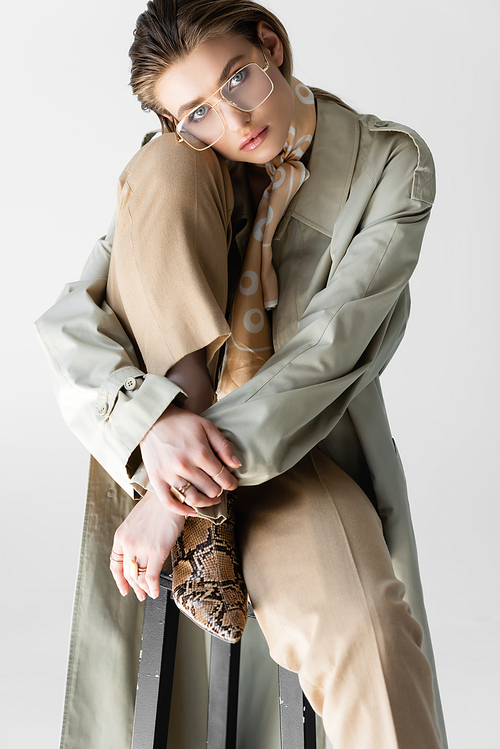 elegant young woman in trench coat, glasses and scarf sitting on chair isolated on white