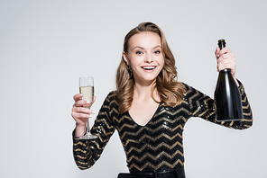 cheerful woman holding glass of champagne and bottle isolated on grey