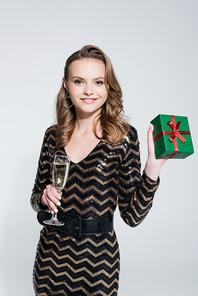 happy young woman holding glass of champagne and wrapped christmas present isolated on grey